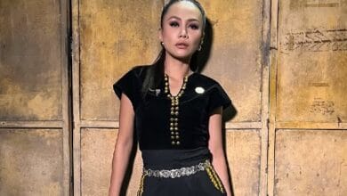 stacy 2 - Wilayah.com.my Woman in a black outfit with gold jewelry standing against a rustic wooden door backdrop.