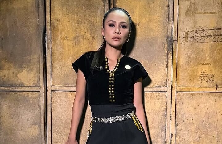 Woman in a black outfit with gold jewelry standing against a rustic wooden door backdrop.
