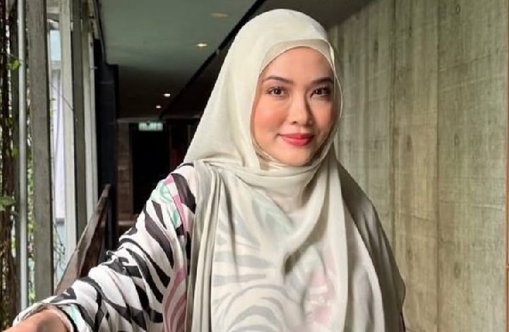 Woman wearing a light hijab and striped outer garment, smiling in a bright hallway.
