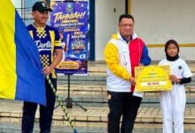 syed faizuddin - Wilayah.com.my Man in a colorful jacket hands a large ceremonial check to a girl in white on school steps; a poster and flag flank them in the background.