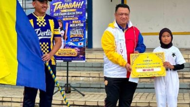 syed faizuddin - Wilayah.com.my Man in a colorful jacket hands a large ceremonial check to a girl in white on school steps; a poster and flag flank them in the background.