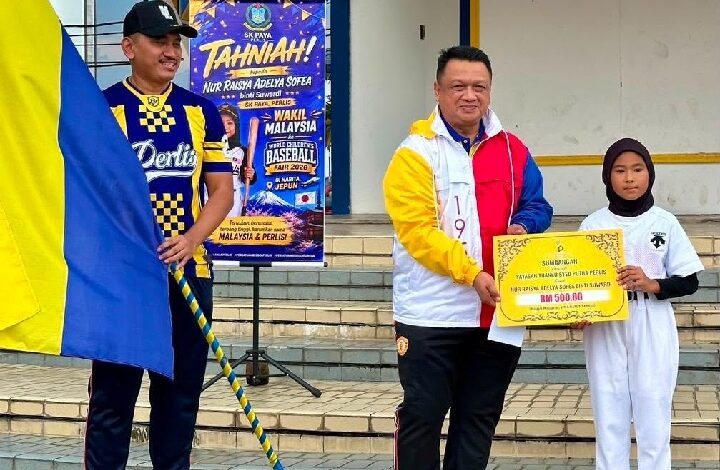 syed faizuddin - Wilayah.com.my Man in a colorful jacket hands a large ceremonial check to a girl in white on school steps; a poster and flag flank them in the background.