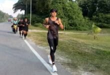syed saddiq - Wilayah.com.my A man running along a road in a black athletic outfit, with a group of runners and a motorcycle behind him on the left, in a green, leafy area.