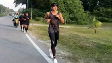 syed saddiq - Wilayah.com.my A man running along a road in a black athletic outfit, with a group of runners and a motorcycle behind him on the left, in a green, leafy area.