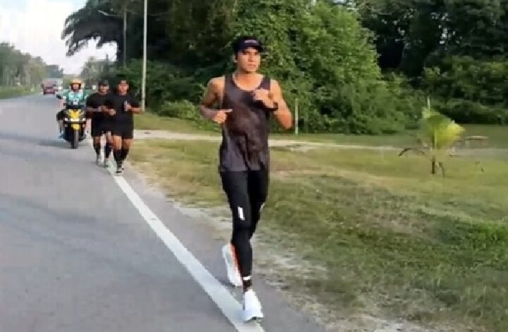 syed saddiq - Wilayah.com.my A man running along a road in a black athletic outfit, with a group of runners and a motorcycle behind him on the left, in a green, leafy area.