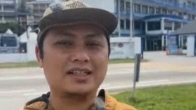 tamim dahri - Wilayah.com.my Man wearing a tan cap and yellow jacket speaks to the camera outdoors, with a road and blue buildings in the background.