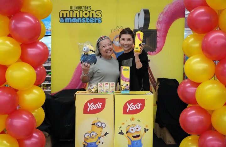yeos - Wilayah.com.my Two women stand behind a yellow Minions & Monsters promotional booth with balloons on either side, smiling at the camera.