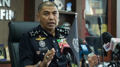 High-ranking police officer in uniform speaks at a press conference, gesturing with hands, with microphones in front and awards/display case in the background.